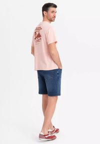 Camiseta de algodón rosa claro con diseño gráfico en la parte trasera. Combinada con pantalones cortos de mezclilla oscura y zapatillas deportivas rojas, blancas y grises.