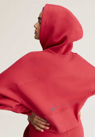 Person trägt einen leuchtend roten Kapuzenpullover, steht im Profil mit den Händen in den Hüften und zeigt Rück- und Seitendetails des Kleidungsstücks.