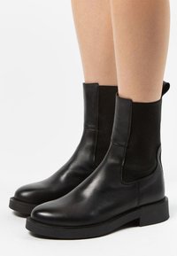 Bottines noires en cuir lisse avec des panneaux latéraux élastiques, un bout rond et un talon plat, présentant des détails de couture sur le corps.