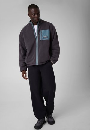 Homme portant une veste polaire foncée avec une fermeture éclair bleue et une poche, un pantalon noir et des chaussures noires, debout devant un fond gris uni.
