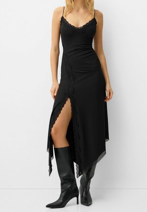 Femme portant une robe noire sans manches avec une bordure en dentelle, une fente haute sur la cuisse et des bottes noires à talons montant jusqu'aux genoux, debout devant un fond blanc.