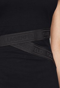 Černý bavlněný top s širokým matným černým elastickým pásem s vyraženým nápisem "KARL LAGERFELD", který protíná přední část oděvu.