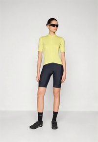 Femme debout dans un maillot de cyclisme jaune à manches courtes, un short rembourré noir, des chaussettes noires, des chaussures de cyclisme et de grandes lunettes de soleil.