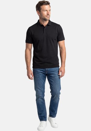 Uomo in piedi che indossa una polo nera a maniche corte, jeans blu e sneakers bianche, che guarda di lato contro uno sfondo neutro.