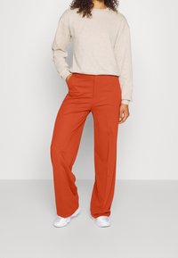 Červené kalhoty se širokými nohavicemi s hladkým povrchem, spárované s krémovým svetrem. Oblečení doplňují bílé tenisky.