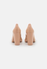 Chaussures à talons hauts en cuir verni nude avec bout pointu et talon bloc, présentant une finition lisse et brillante ainsi qu'un design minimaliste.