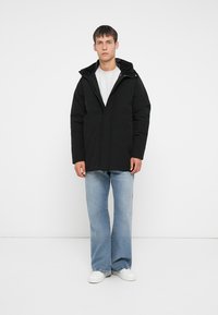 Svart isolert jakke med hette, glidelås og sidelommer. Brukt over en lys grå hettegenser og kombinert med lyseblå jeans.
