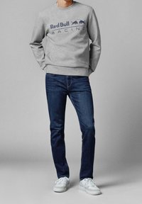 Szara bluza z kapturem z logo "Red Bull Racing" i niebieskimi napisami, zestawiona z ciemnoniebieskimi dżinsami i białymi trampkami. Prosty, swobodny design.