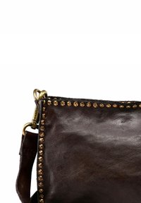 Angolo di una borsa a mano in pelle marrone scuro con dettagli in ottone, e un clasp in ottone che attacca la tracolla alla borsa.