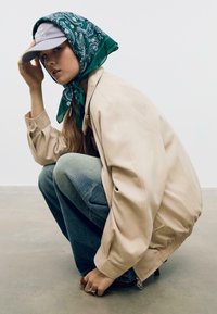 Béžová oversized bunda s elastickým spodným lemom, skombinovaná s modrými džínami. Zelený šatka s bielym paisley vzorom a šedá čiapka dotvárajú vzhľad.