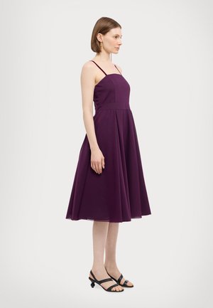 Purple midi dress with thin straps, fitted bodice, and flared skirt. Smooth texture, minimalistic design. Black strappy shoes complement the outfit.