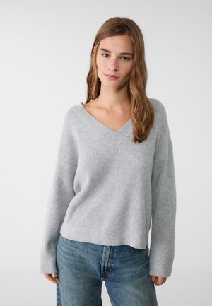 Grijze gebreide trui met een V-hals, lange mouwen en een relaxte pasvorm. De stof heeft een ribbelstructuur en de onderkant is iets verkort. Draagt comfortabel met blauwe spijkerbroeken.