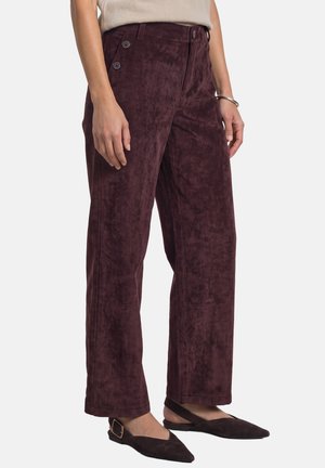 Femme portant un pantalon en velours côtelé violet foncé, ample, avec des boutons sur le côté, un haut beige et des chaussures plates noires pointues avec des brides à boucle.