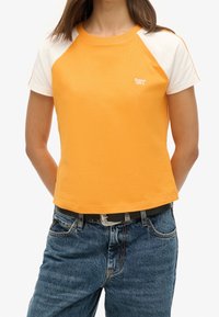 Camiseta de algodón naranja con mangas raglán blancas. Presenta un escote redondo y un pequeño logo en el pecho. Diseño corto y ajustado.
