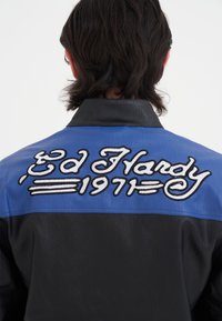 Läderjacka med svart kropp och blå axelpanel. Baksidan har vit broderad text "Ed Hardy 1971" med dekorativa detaljer.