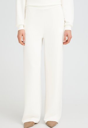 Person trägt weiße, hoch taillierte, weit geschnittene Hose kombiniert mit hellbraunen spitzen Schuhen und steht vor einem schlichten hellen Hintergrund.