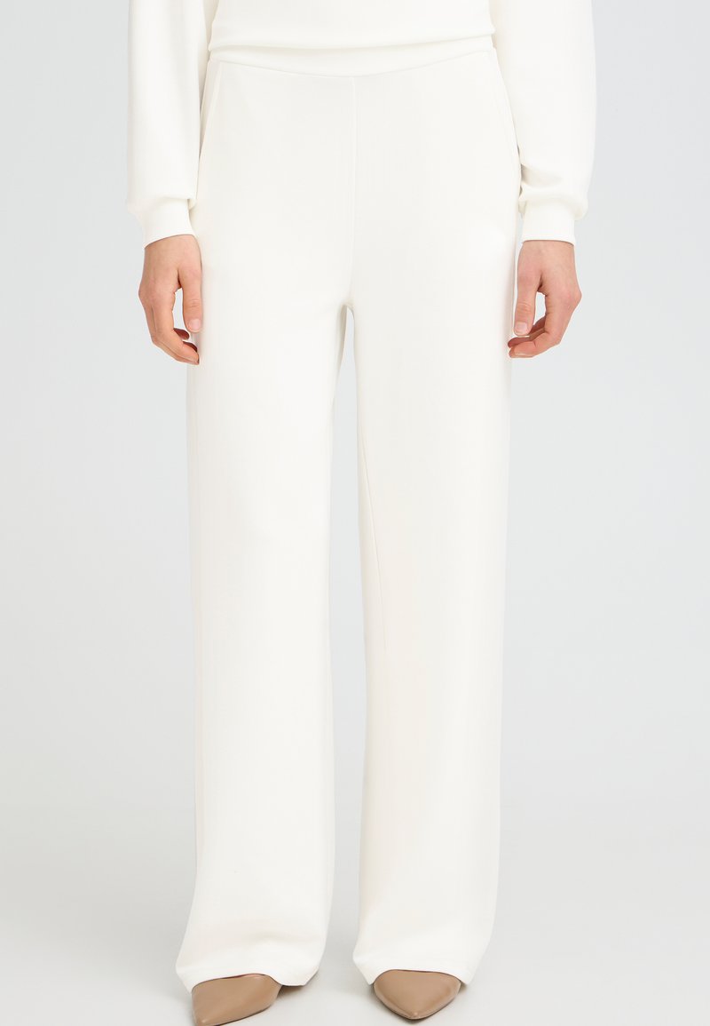 Henkilö pukeutuneena valkoisiin, korkeavyötäröisiin ja leveälahkeisiin housuihin, yhdistettynä ruskeisiin, teräväkärkisiin kenkiin, seisomassa yksinkertaisen vaalean taustan edessä.