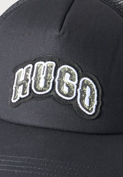 Черен бейзболен шапка с извит периферия, с издигнато лого с надпис "HUGO" в бели контурирани камуфлажни букви.
