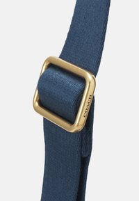 Bracelet en tissu bleu passé à travers une boucle carrée dorée gravée du nom de la marque Coach sur un côté.