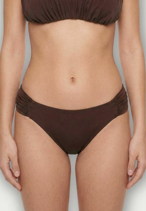 Femme portant un bas de bikini marron avec des côtés froncés, montrant la taille et les mains détendues sur les côtés, devant un fond uni.