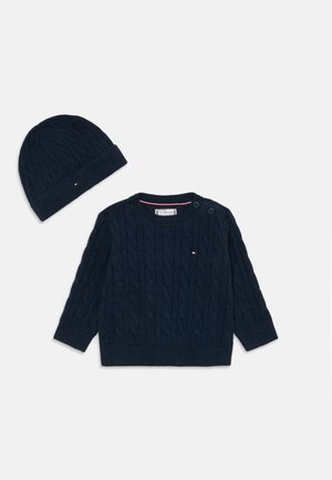 Suéter em azul-marinho com tricot de cabos, com botões no ombro e bainha canelada, combinado com um gorro de malha correspondente.
