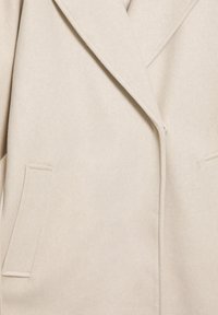Manteau beige sur mesure en mélange de laine doux et texturé, avec un col cranté, une fermeture à un bouton et deux poches avant.
