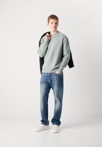 Hellgrüner gerippter Pullover, blaue Jeans, weiße Sneakers. Das Model hält eine schwarze Jacke über der Schulter und steht vor einem neutralen Hintergrund.