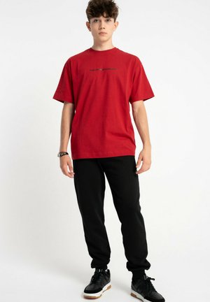 Młody mężczyzna stoi, ubrany w czerwoną koszulkę z grafiką, czarne dresowe spodnie, czarne sneakersy i zegarek na czarnej białym tle.