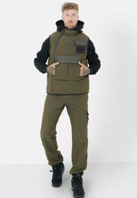 Chaleco acolchado verde oliva sobre una sudadera negra con capucha, con múltiples bolsillos frontales y cierre de cremallera, combinado con pantalones cargo a juego y zapatillas negras.