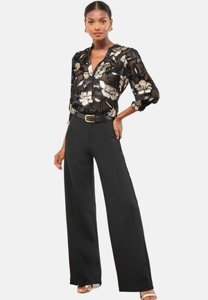 Black sheer blouse with floral design, paired with high-waisted black wide-leg trousers and a gold buckle belt. Model wearing metallic sandals.