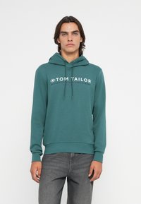 Felpa con cappuccio verde acqua con logo anteriore bianco, dettagli con cordoncino e polsini e orlo a coste, abbinata a jeans grigi scuri.