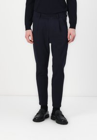 Navy pleated trousers made of fabric with a smooth texture, featuring a button closure and tapered legs, paired with black loafers.