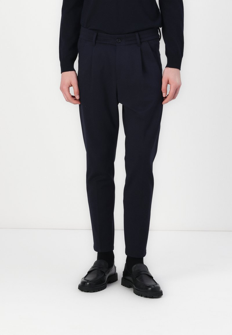 Navy pleated trousers made of fabric with a smooth texture, featuring a button closure and tapered legs, paired with black loafers.