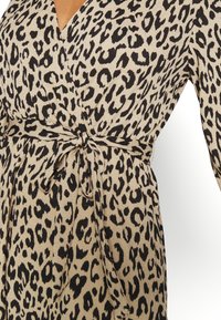 Šaty s leopardím potiskem ve světlé béžové barvě; mají hluboký V-výstřih, dlouhé rukávy a zavázaný pas pro přiléhavý siluetu.