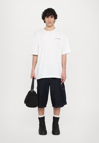 T-shirt blanc à manches courtes avec texte noir, associé à un short cargo en denim foncé avec poches latérales, sac noir et bottes noires zippées.