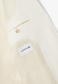 Interno di una giacca color crema che mostra una tasca con bottone e un'etichetta bianca con testo nero "LACOSTE" e un logo a forma di coccodrillo verde.