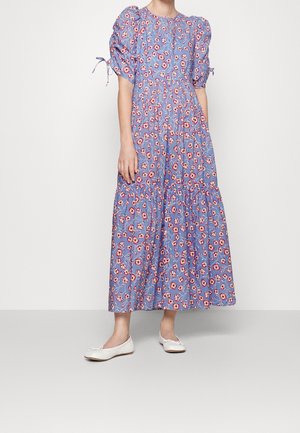 Kobieta w niebieskiej, kwiecistej sukience maxi z bufkowymi rękawami związanymi na łokciach, ubrana w białe baletki, na jednolitym tle.