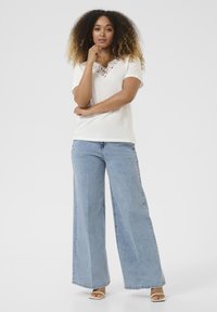 Witte top met korte mouwen en een kanten detail bij de halslijn, gecombineerd met losse lichtblauwe denim wijde broek en beige open schoenen.