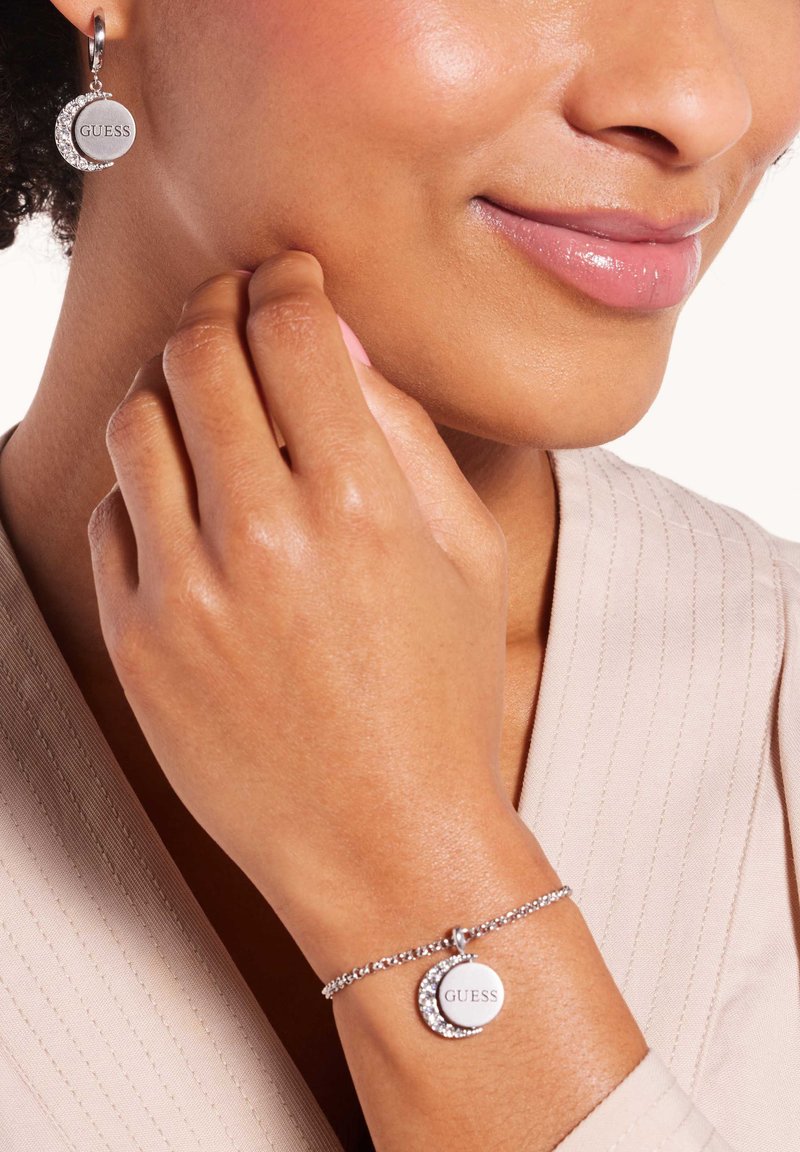 Zilveren sieradenset met cirkelvormige oorbellen en een armband, beide met een gladde afwerking en "GUESS"-merk, aangevuld met strass accentueringen.