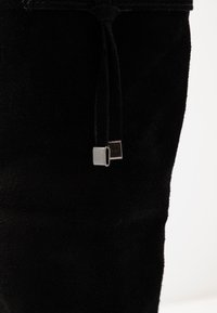 Primer plano del eje de una bota de ante negra con dos cordones negros que terminan en puntas metálicas rectangulares de plata.