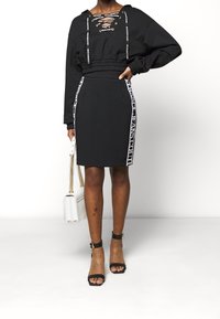 Svart avklippt sweatshirt med snörning framtill, i kombination med en svart pennkjol med vit sidobranding. Modellen håller en vit handväska.