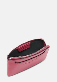 Pochette en cuir rose avec fermeture éclair, bords arrondis, surface texturée et intérieur en tissu noir. Présente un petit logo en relief à l'intérieur.