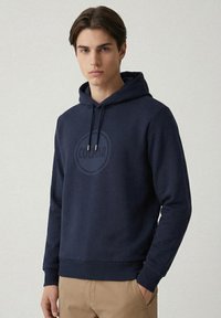 Felpa con cappuccio in cotone blu navy con stampa di un logo rotondo al centro, cordini regolabili, polsini e orlo a coste. Il modello indossa pantaloni kaki.
