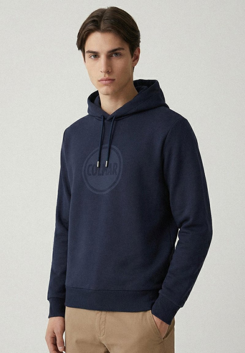 Felpa con cappuccio in cotone blu navy con stampa di un logo rotondo al centro, cordini regolabili, polsini e orlo a coste. Il modello indossa pantaloni kaki.