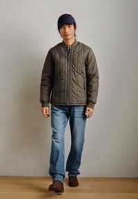 Jachetă căptușită de culoare verde măslinie, cu fermoar frontal, asortată cu blugi ușor albaștri și pantofi maro. Actorul poartă o șapcă navy. Fundal simplu.