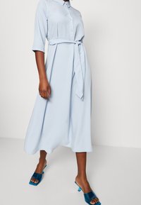Abito midi azzurro chiaro con apertura frontale a bottoni, colletto e vita segnato da una cintura, abbinato a mule aperti turchesi e texture del tessuto liscia.