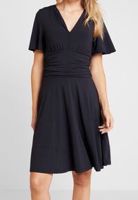 Svart klänning i mjuk tyg, med V-ringning, korta fladdrande ärmar, drapering vid midjan och en utsvängd kjol.