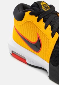 Gul og sort atletisk sko med netoverdel, iøjnefaldende rød accent og en fremtrædende sort Nike-swoosh. Har en hvid mellemsål og teksturerede detaljer.