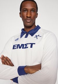 Baltas polo marškinėlis ilgomis rankovėmis su tamsiai mėlyna apykakle; ant krūtinės parašyta didelėmis tamsiai mėlynomis raidėmis „EMST“, viršutiniame kairiajame kampe yra logotipas. Audinys yra tekstūruotas, rankogaliai mėgintuvėlio tipo.