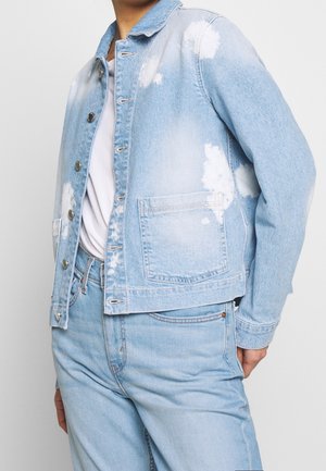 Chaqueta vaquera de mezclilla azul claro con manchas blancas de lejía, usada sobre una camisa blanca, combinada con jeans de mezclilla azul claro a juego.